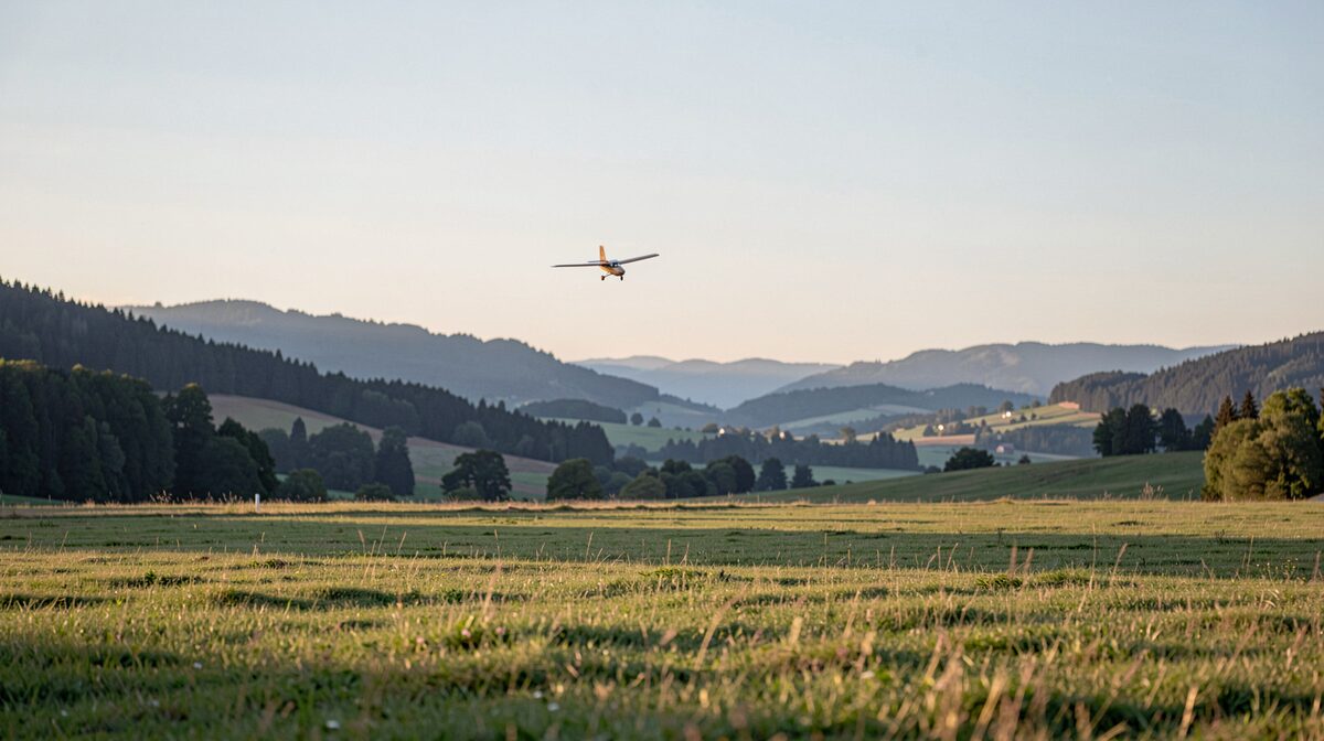 A grass flying field at the edge of the Swabian Alb hills in late afternoon light, a small model glider rising against the sky, wide landscape framing.